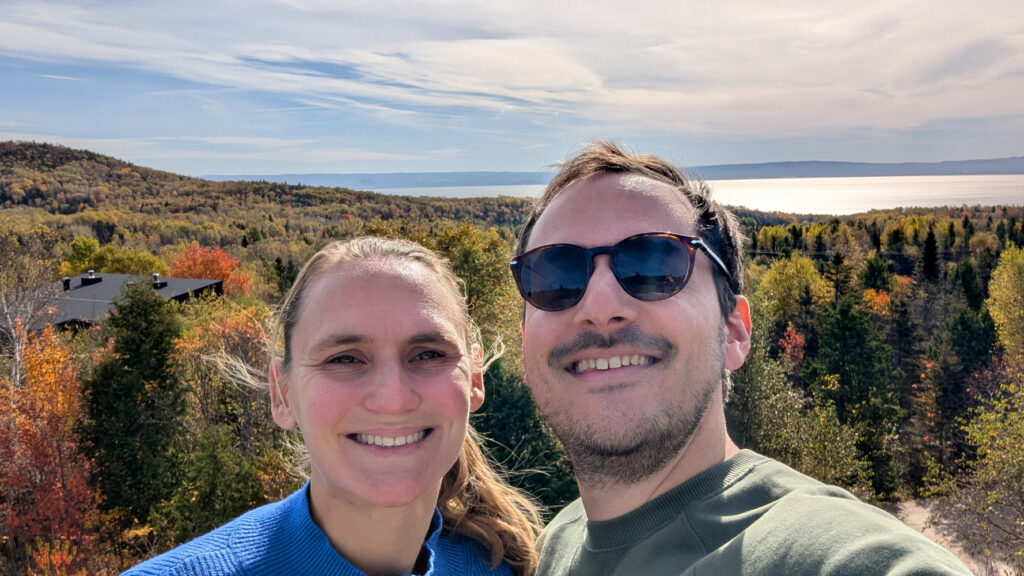 Un couple souriant se prend en selfie sur une colline boisée en automne, avec un grand lac, des montagnes lointaines et un ciel lumineux au loin.