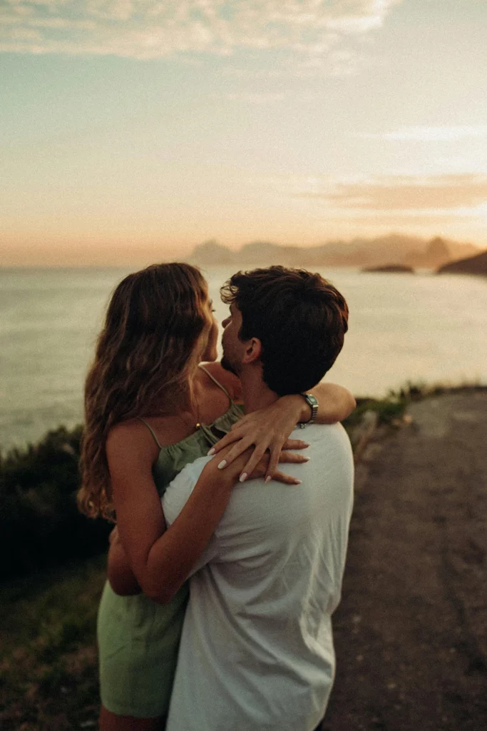Au coucher du soleil, un couple s’étreint face à la mer sur une côte rocheuse, dans une ambiance paisible de voyage sensoriel et romantique.