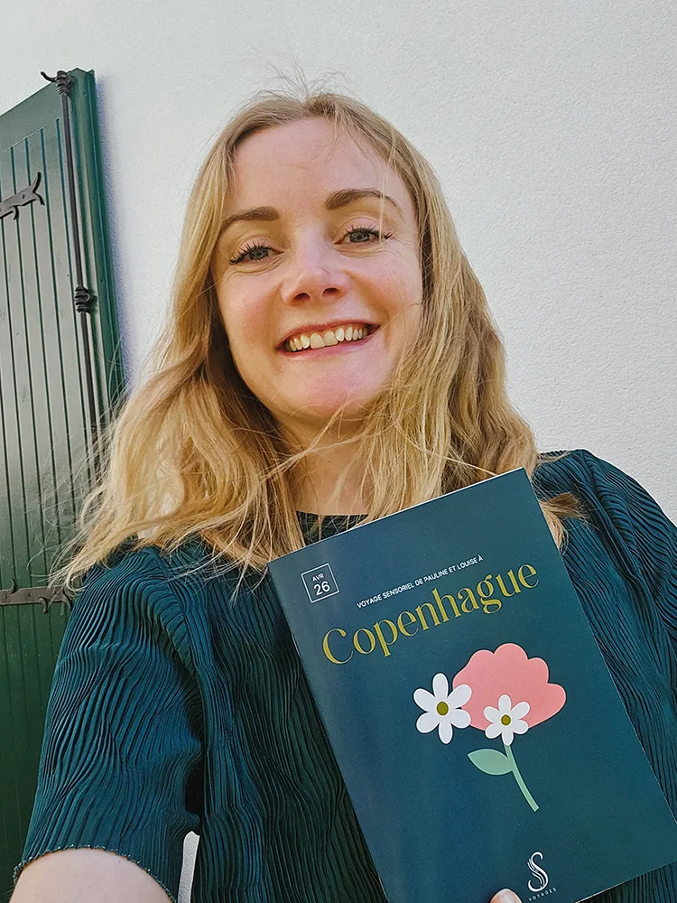 Florence sourit en tenant un carnet Copenhague signé Sillage Voyages, dans un univers de voyage sensoriel inspiré par Fragonard et les parfums.