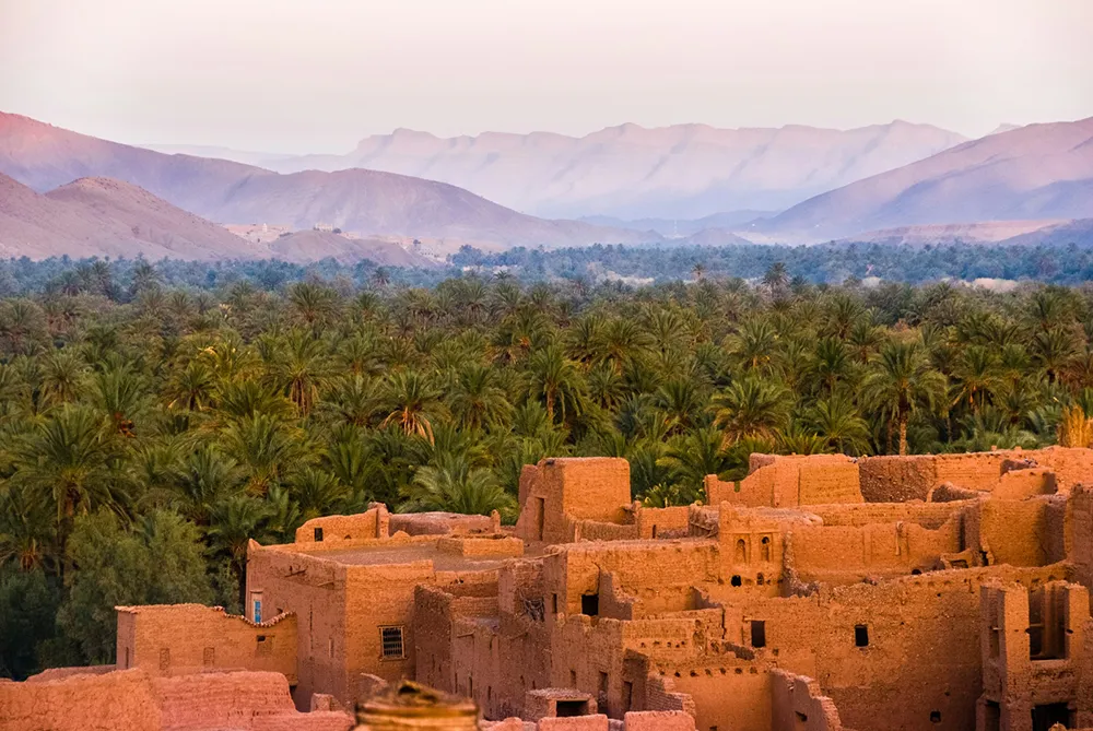 Au Maroc, un ksar en terre ocre domine une palmeraie verdoyante, avec les montagnes de l’Atlas en arrière-plan sous une lumière douce.