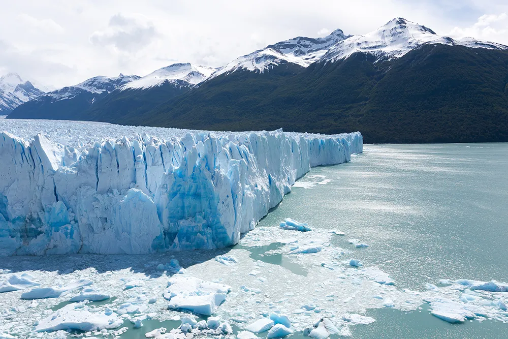 Un immense glacier bleu avance dans une baie de Patagonie, au pied de montagnes enneigées et d’eaux glacées en Argentine, au sud.